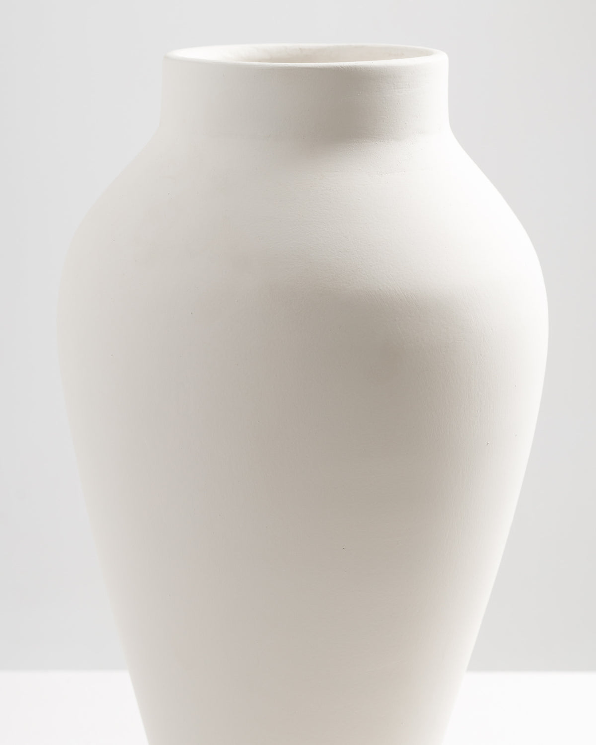 A smooth, white ceramic vase with an elegant, curvy silhouette sits against a light background. The vase features a narrow opening at the top and a rounded body, showcasing a minimalist design that highlights its simplicity.