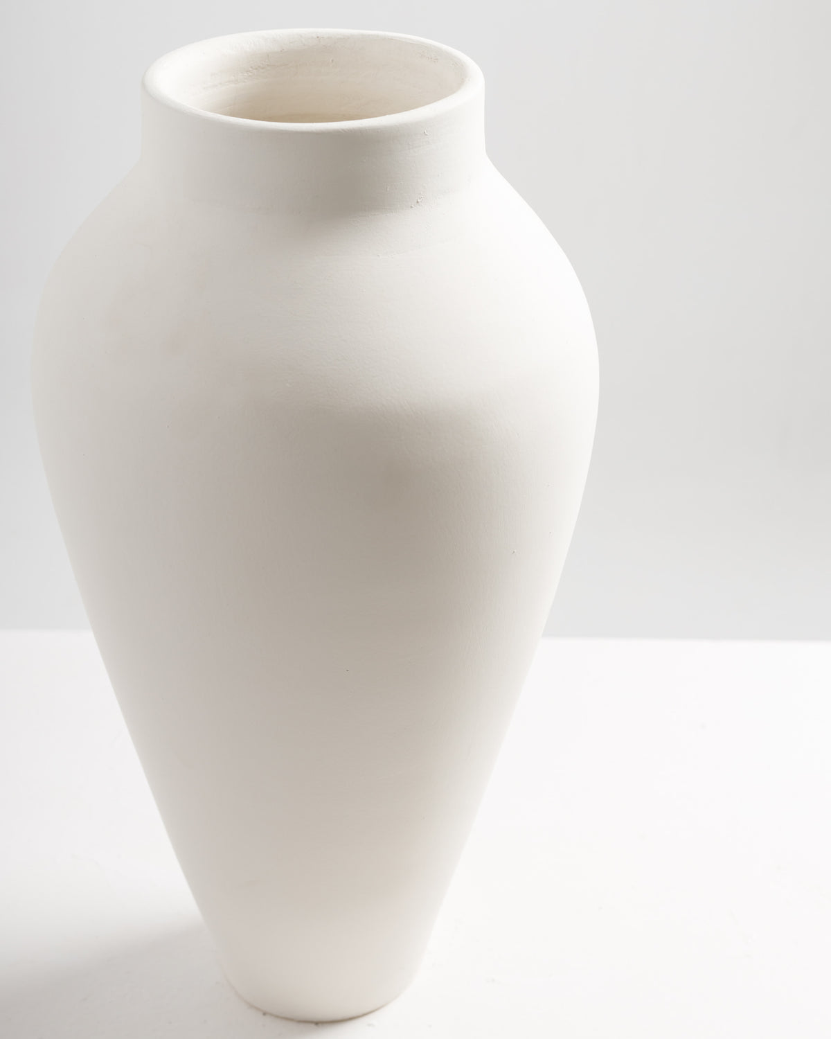 A tall, elegant vase with a smooth, matte white finish stands upright against a light background. The vase features a wide body that tapers towards the base, creating a classic silhouette perfect for displaying flowers or as.