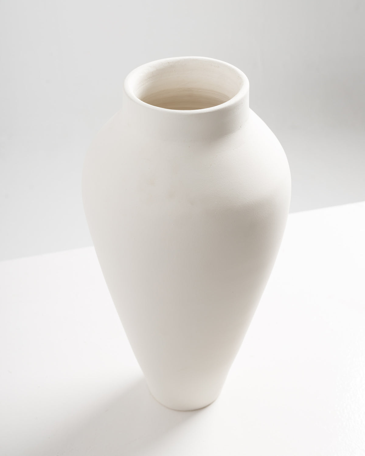 A simple, elegant white vase with a smooth, matte finish and a slender, elongated shape. The vase features a wide mouth and tapers towards the base, set against a minimalist background that enhances its modern aesthetic.