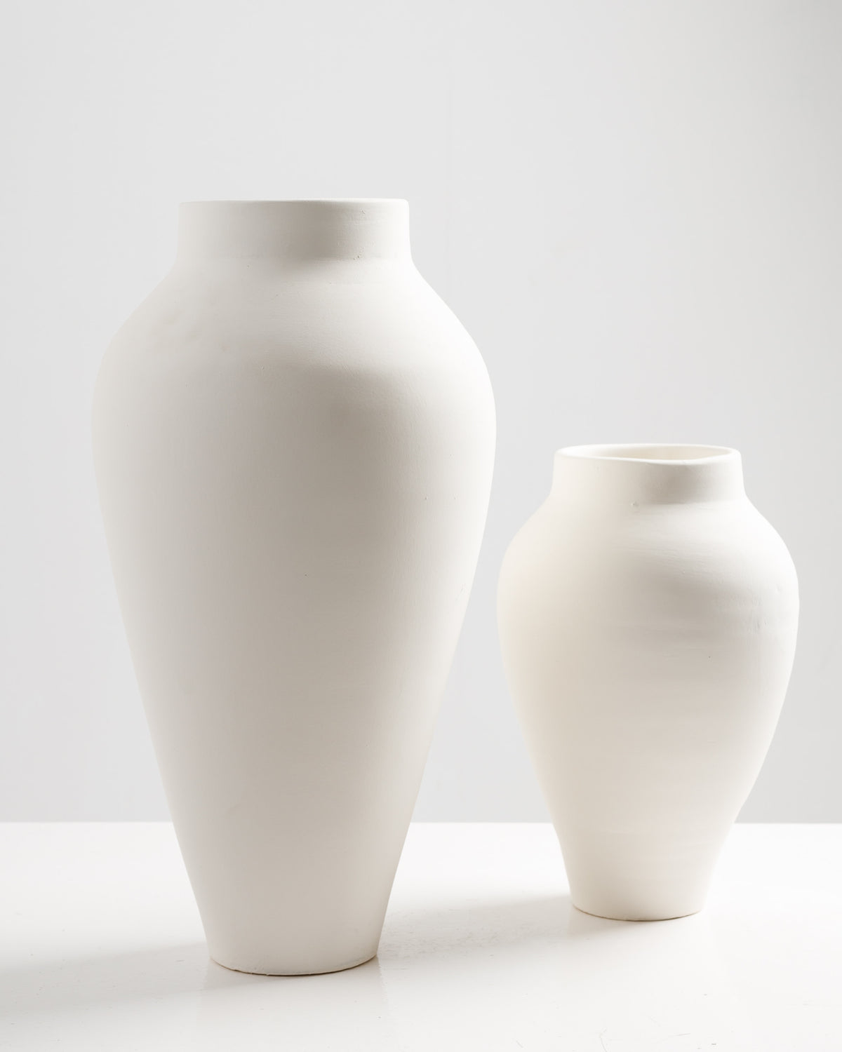 Two elegant, minimalist ceramic vases in a matte white finish are displayed side by side. One vase is significantly larger, with a rounded body and a wide neck, while the smaller vase features a more slender silhouette.