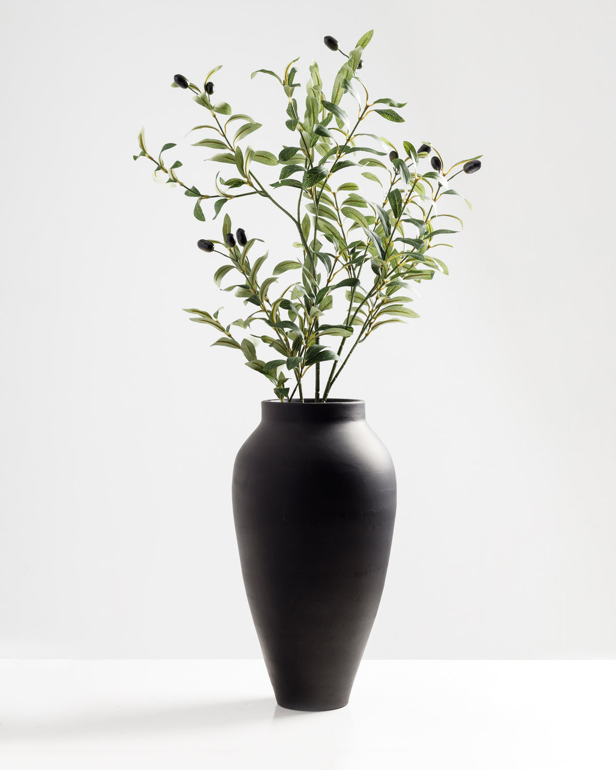 A tall, matte black vase holds a decorative arrangement of artificial olive branches featuring silk leaves and foam fruits. The natural-looking greenery adds an elegant touch to any space, requiring no maintenance.