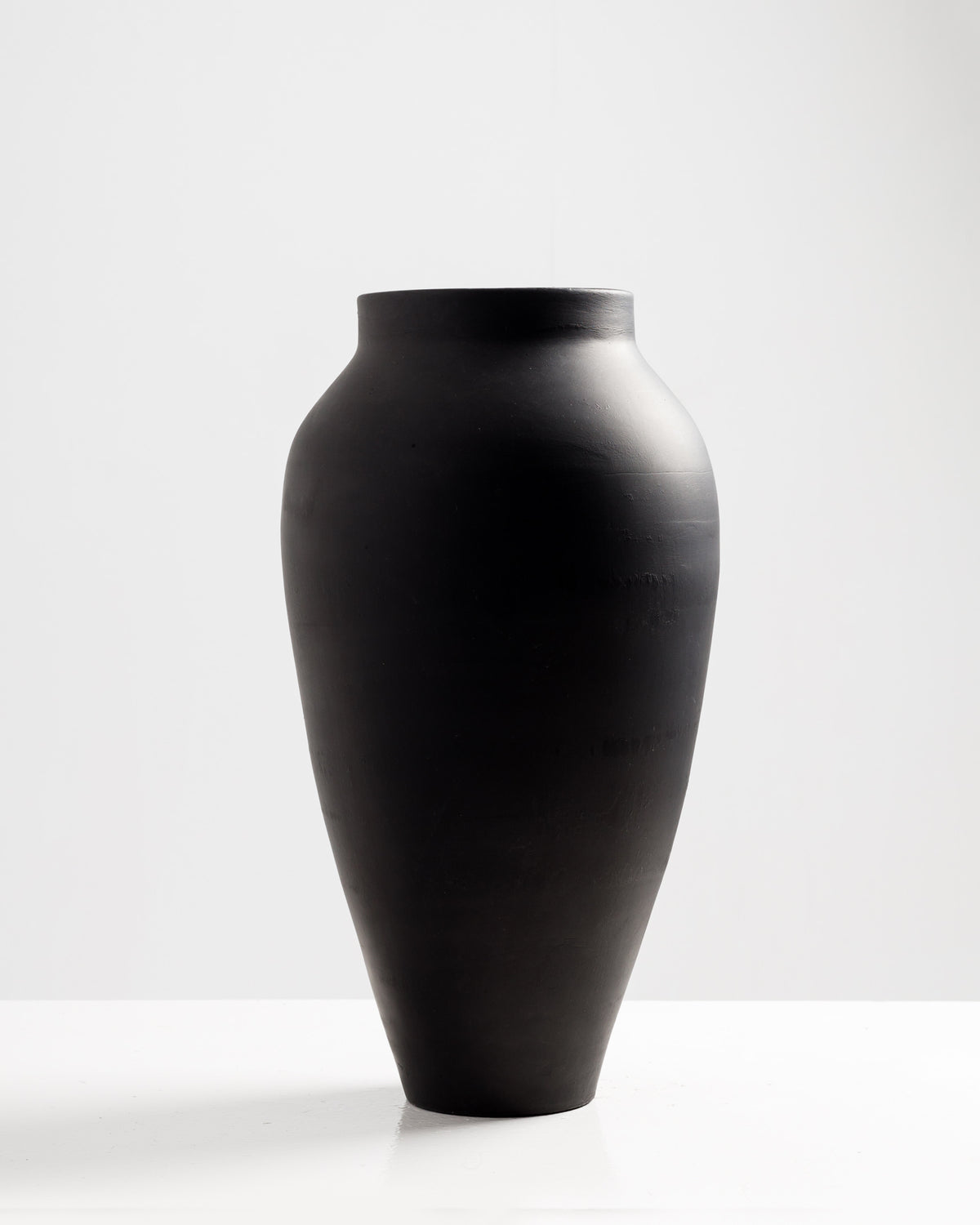 A tall, sleek black vase with a smooth, rounded body and a narrow neck stands on a white surface against a light background. The minimalist design emphasizes its elegant shape, making it a striking decorative piece.