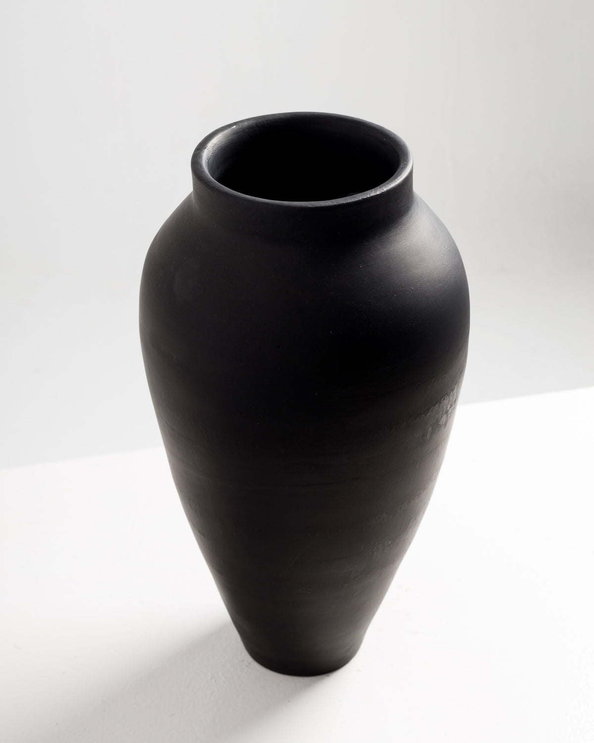A tall, matte black vase with a rounded body and a narrow neck sits on a white surface, showcasing a simple and modern design. The vase features a smooth finish and elegant silhouette.
