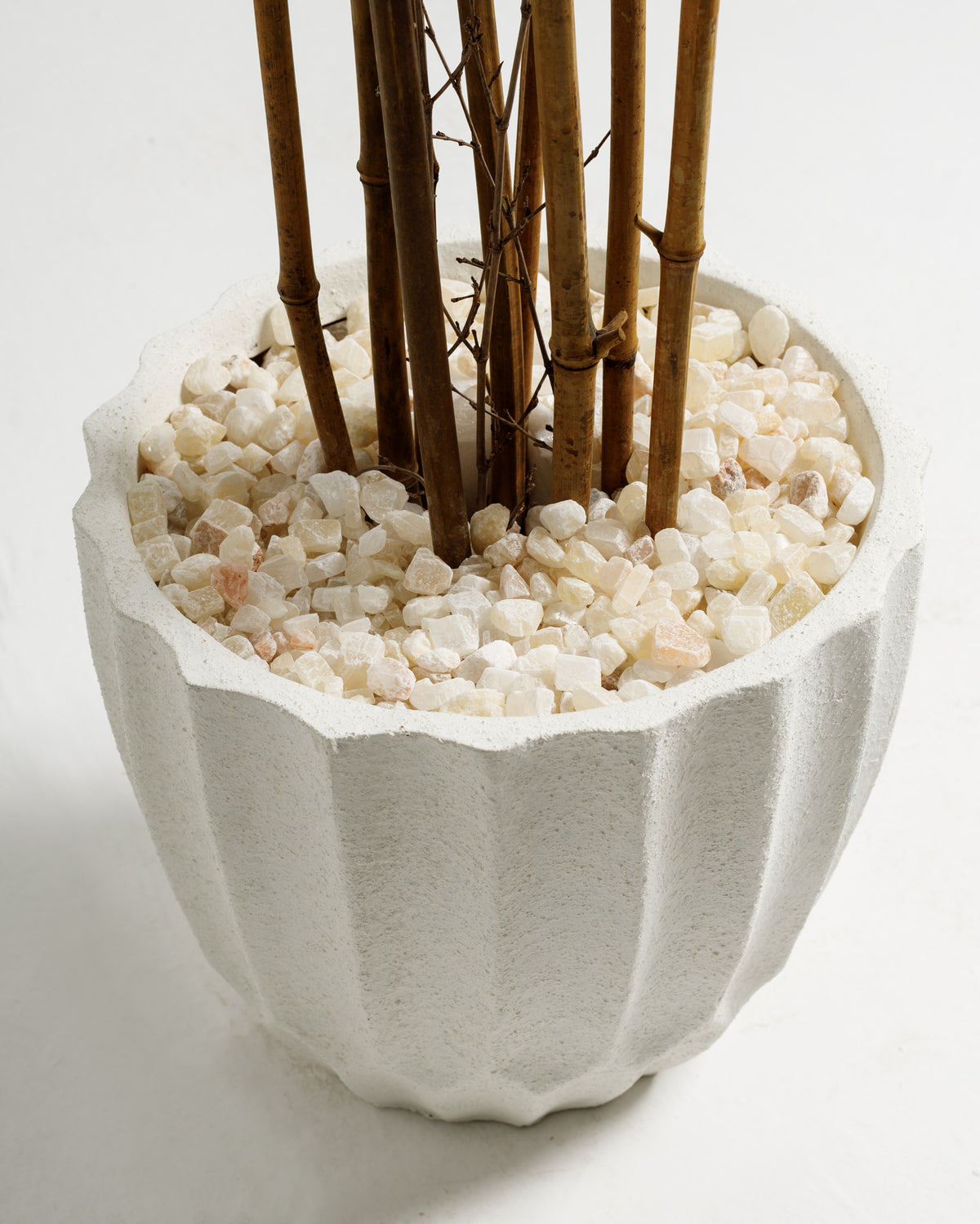 A textured white planter with a fluted design holds several tall, slender bamboo stems. The base is filled with small white and light-colored stones, creating a clean.