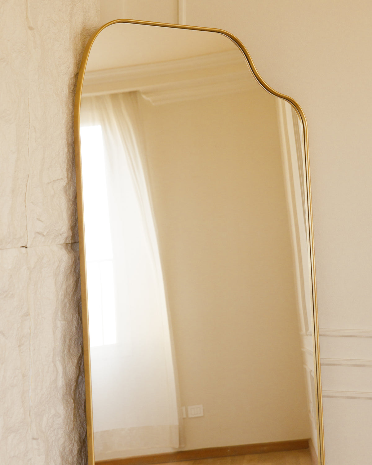 A stylish full-length mirror with a softly curved top and a sleek gold frame, leaning against a textured white wall. Natural light filters in through nearby sheer curtains, enhancing the elegant design and ambiance of the room.