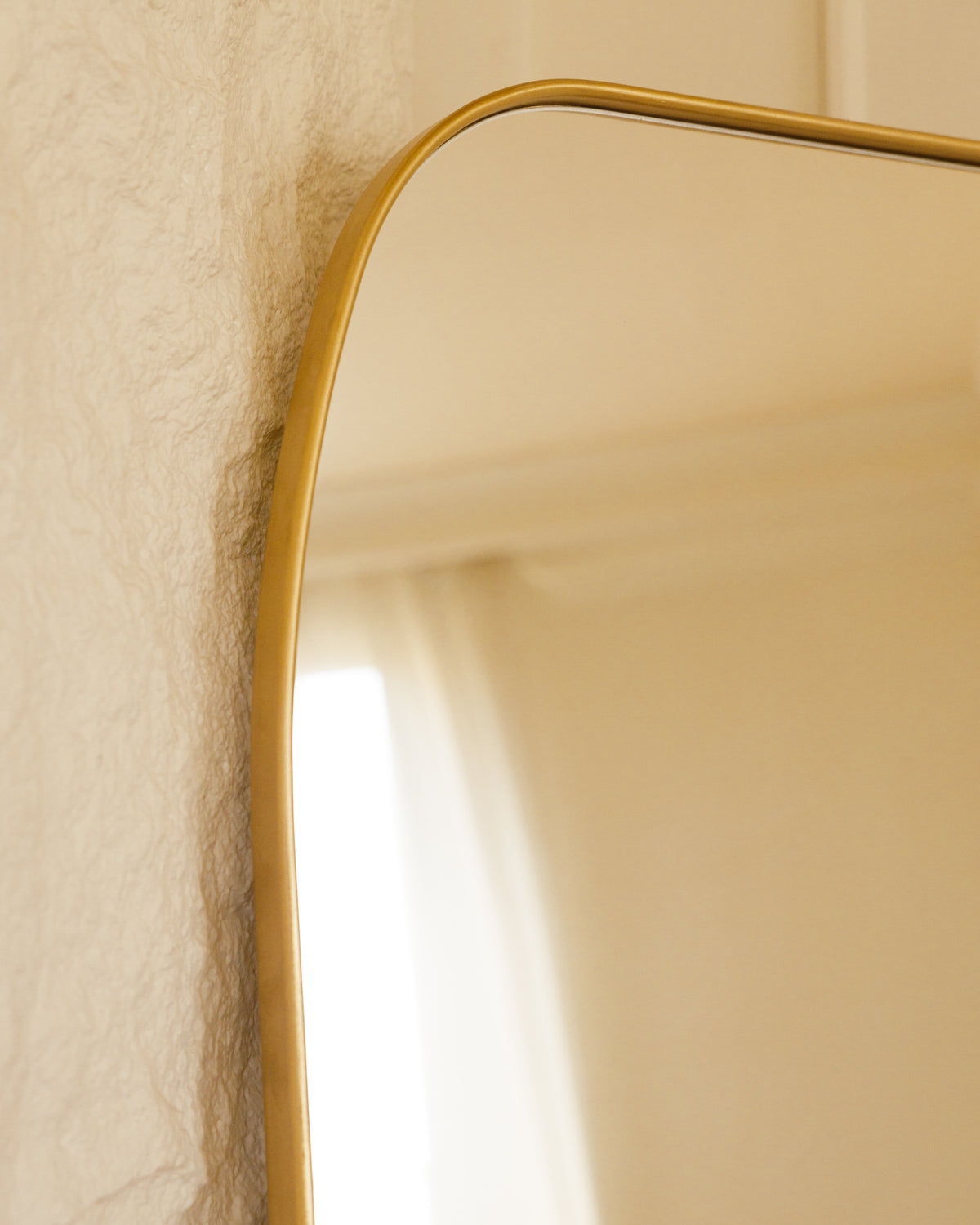 Close-up view of a gold-framed mirror reflecting soft, natural light. The mirror is set against a textured wall, highlighting its elegant curves and modern design, perfect for enhancing any interior space.