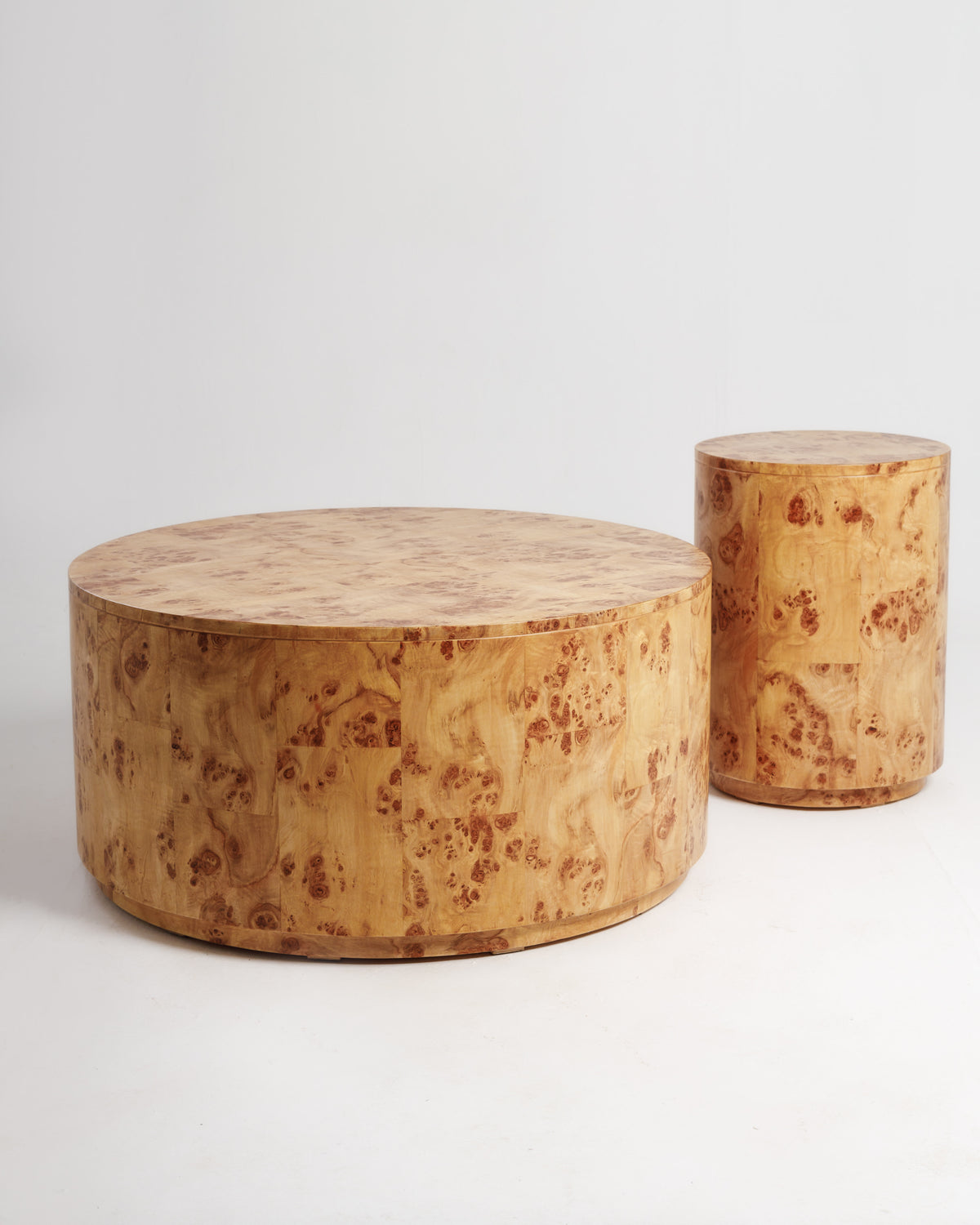 A set of two round tables made from burlwood, featuring a smooth, natural finish. The larger table is low and wide, while the smaller one is cylindrical and slightly taller, showcasing the unique grain patterns of the wood.