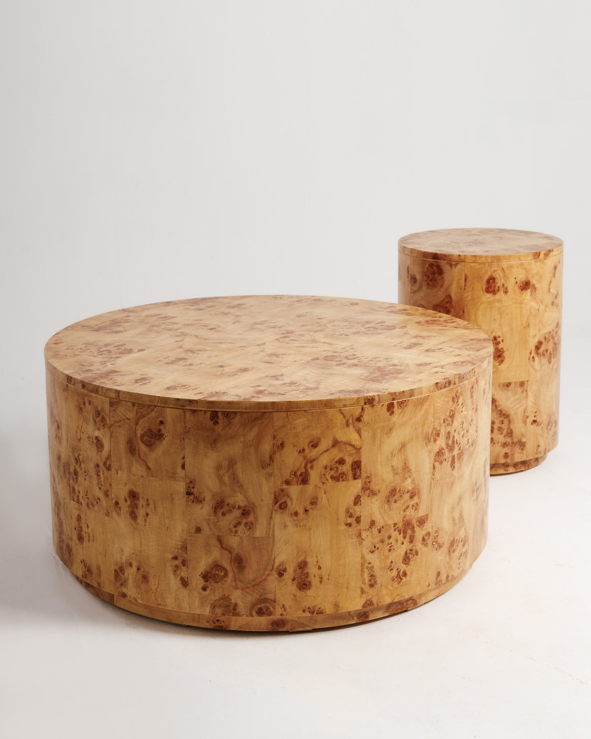A round burlwood coffee table with a smooth, polished surface showcasing intricate wood grain patterns sits prominently in the foreground, accompanied by a smaller cylindrical table nearby.