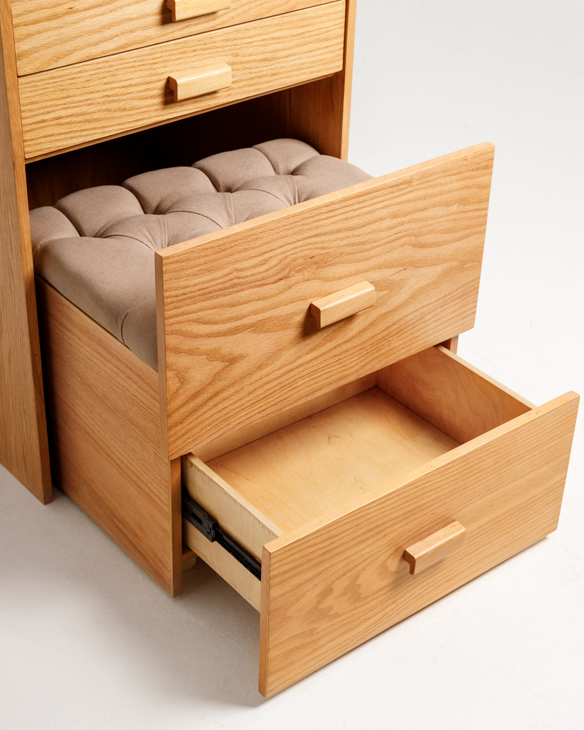 A wooden dresser with a light oak finish features two open drawers, one partially pulled out. The top drawer has a tufted cushioned seat in a neutral color, creating a stylish and functional storage solution for various essentials.