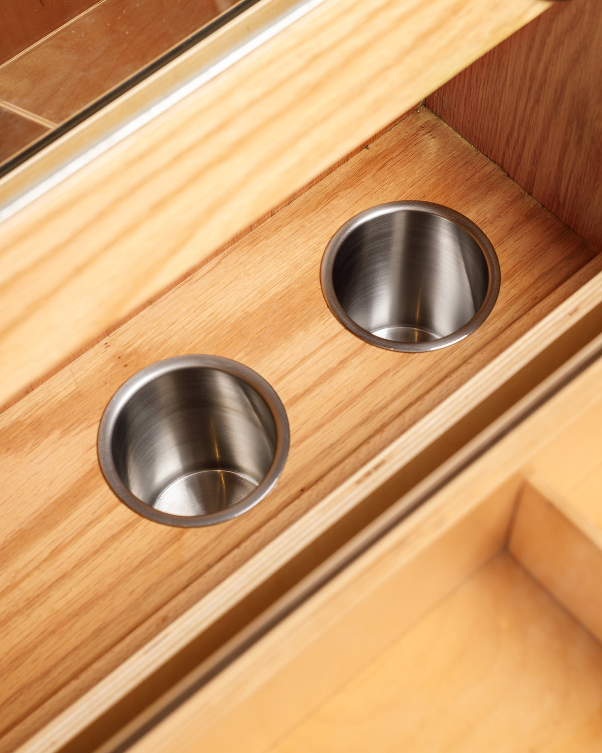 Close-up view of a wooden vanity drawer featuring two circular stainless steel cup holders embedded in the surface. The natural wood finish adds warmth, while the shiny metal accents provide a modern touch.