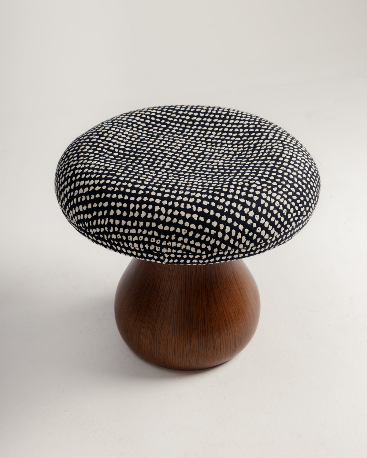 A round pouf features a dark fabric cushion adorned with a white dotted pattern, resting on a smooth, curvaceous wooden base. The design combines modern aesthetics with organic elements, making it a stylish seating option.