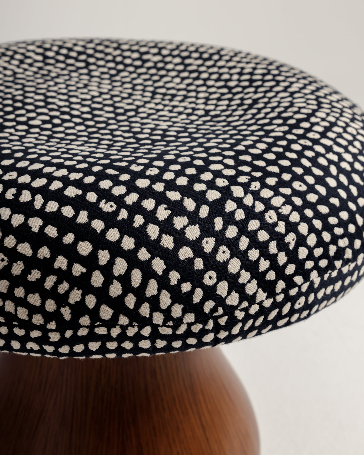 A close-up view of a round pouf featuring a textured fabric surface adorned with a black and cream speckled pattern. The pouf is supported by a smooth, curved wooden base, showcasing a modern and stylish design.