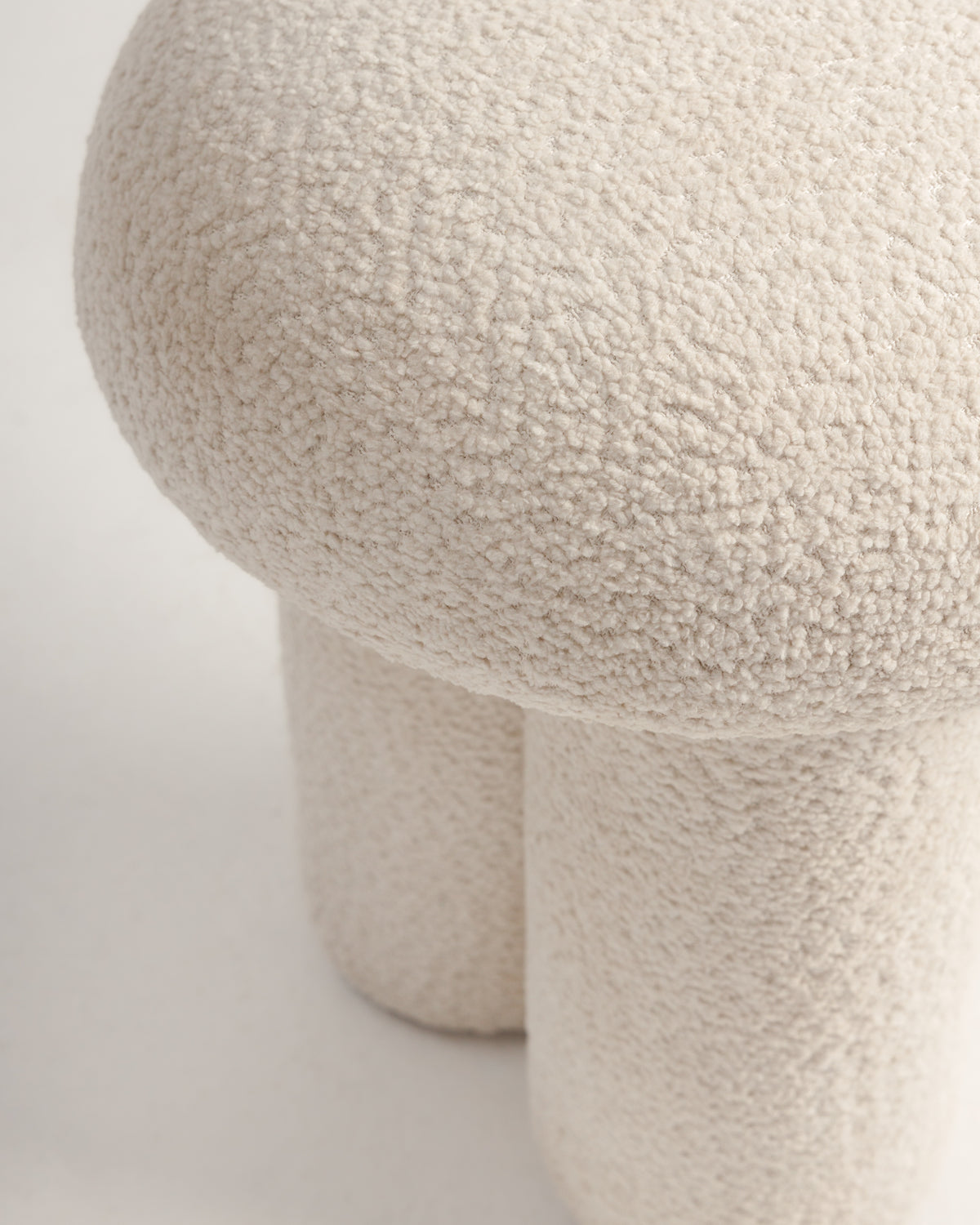 A close-up view of a soft, off-white pouf with a sculptural design. The pouf features a plush, rounded top and a dual cylindrical base, all wrapped in a textured bouclé fabric for a cozy, modern accent.