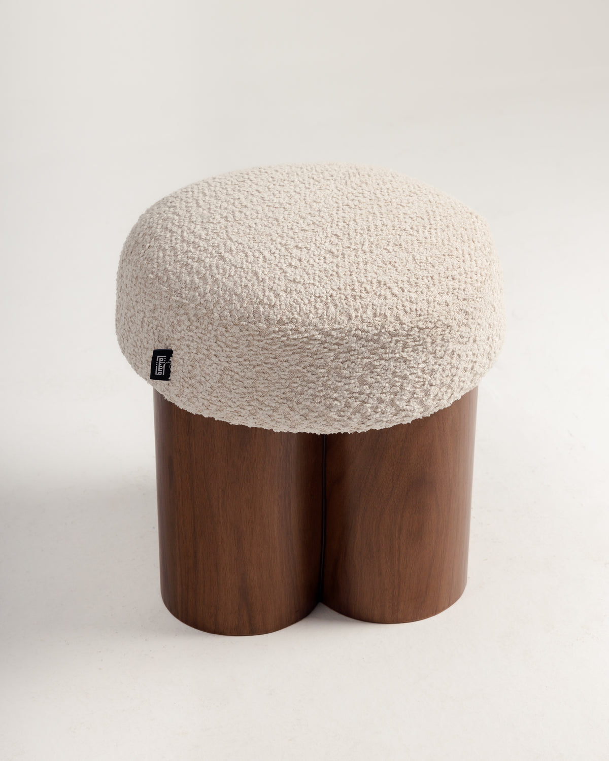 A stylish pouf featuring a plush cream cushion with a textured surface, resting on a unique triple-cylinder walnut wood base. The design combines natural warmth and elegance, making it a versatile accent piece for any space.