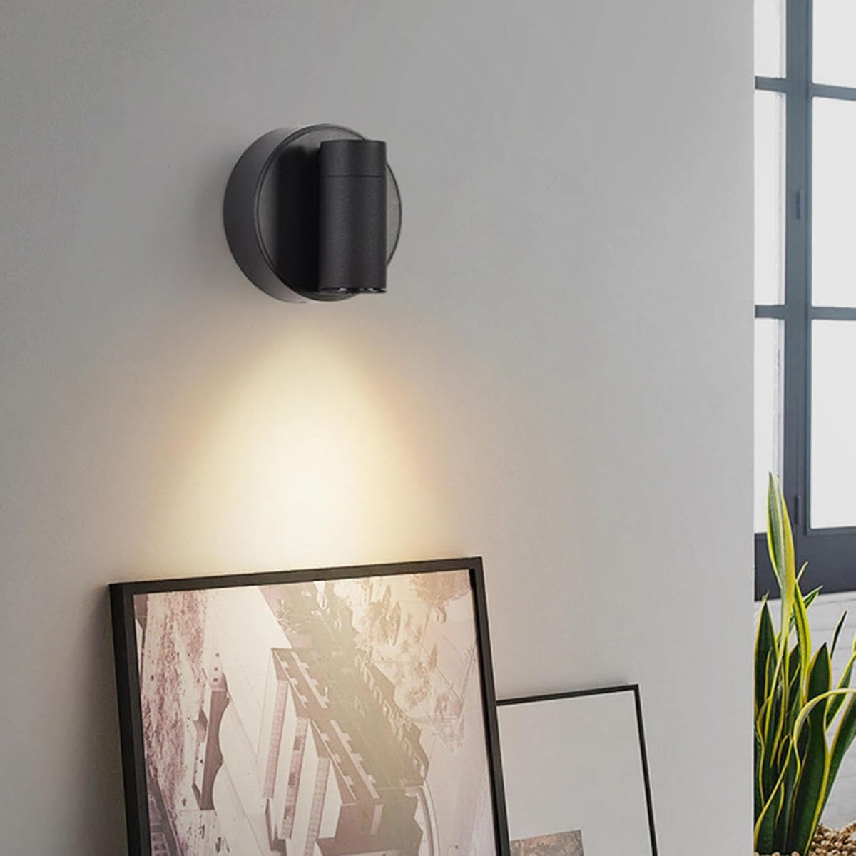 A modern LED wall spotlight is mounted on a light gray wall, emitting a warm glow that illuminates the space below. Two framed pictures are leaning against the wall, and a green plant is visible in the foreground, adding a touch of color.