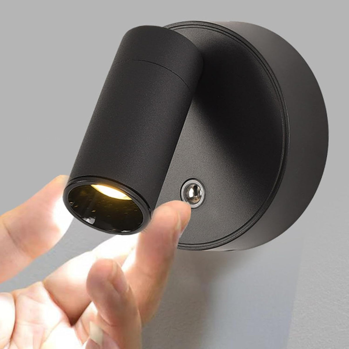 A modern black LED wall spotlight is mounted on a gray wall, with a persons hand reaching toward the light, indicating a touch on/off switch. The spotlight has a cylindrical shape and emits a warm glow, enhancing the contemporary aesthetic.