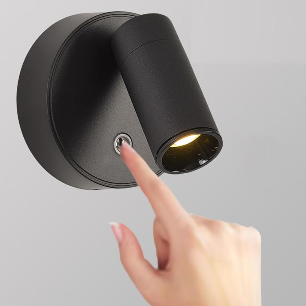 A hand is pressing the touch switch of a modern black LED spotlight mounted on a circular wall plate. The spotlight is angled downward, illuminating a warm light, showcasing its sleek design suitable for contemporary interiors.