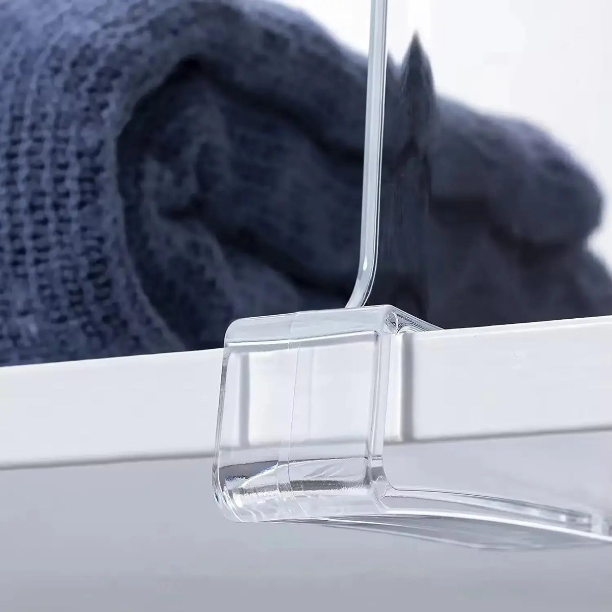 A clear acrylic shelf divider is positioned on a white shelf, securely holding a folded blue sweater next to it. The dividers base is visible, showcasing its design for organizing clothing and maximizing storage space.