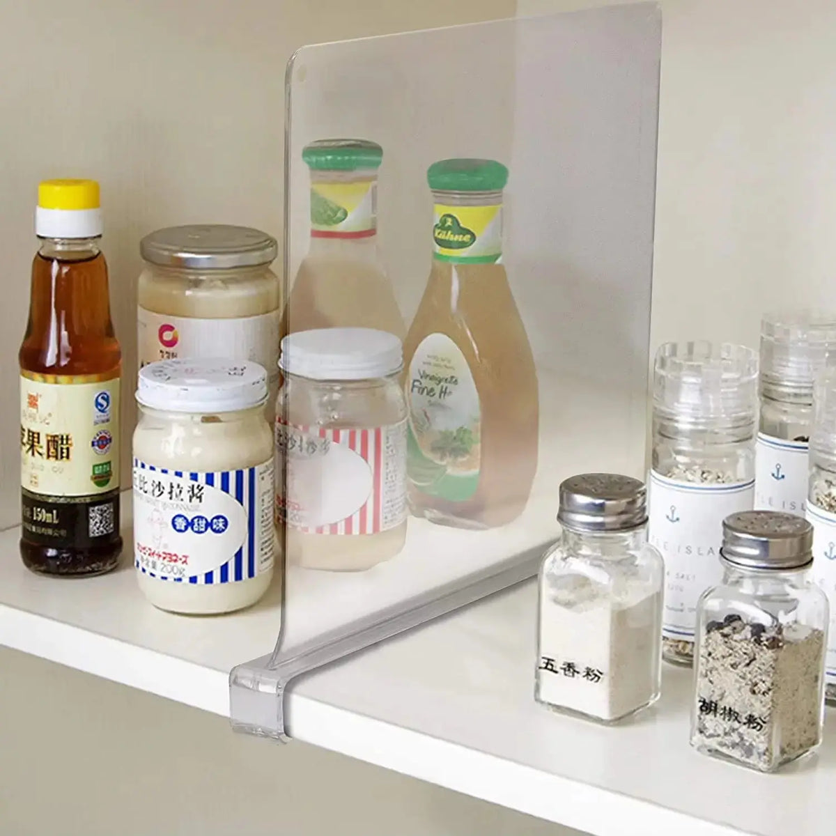 A clear acrylic shelf divider is positioned on a white shelf, separating various jars and bottles. The items include sauces, dressings, and spices, organized neatly behind the divider for easy access and a tidy appearance.